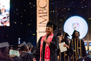 A line of excited graduates wave and smile to the crowd.