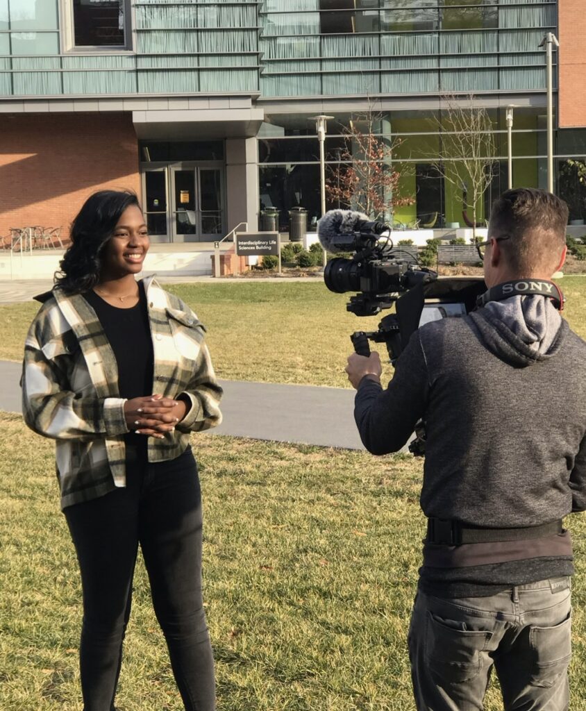 Woman outdoors stands facing a reporter holding a news camera; academic building in background