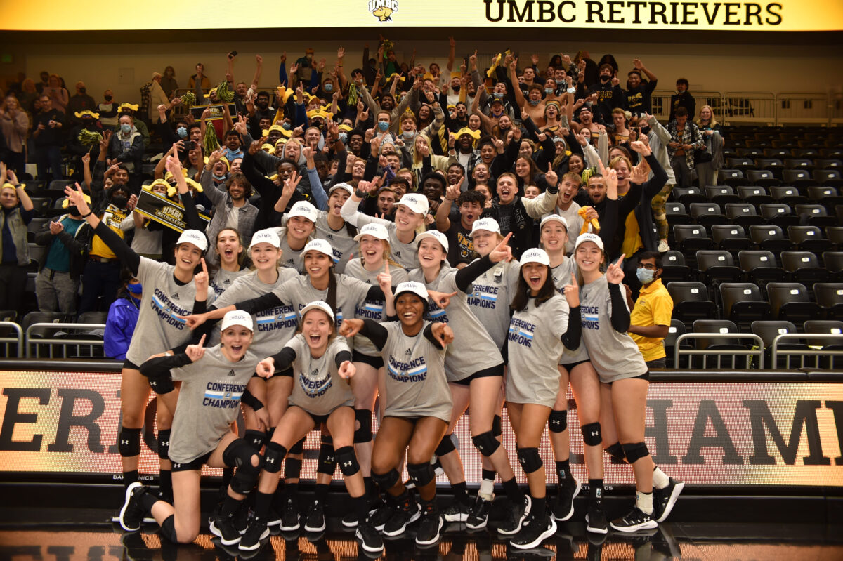 A group of women athletes celebrate. Fans behind them cheer in support. 