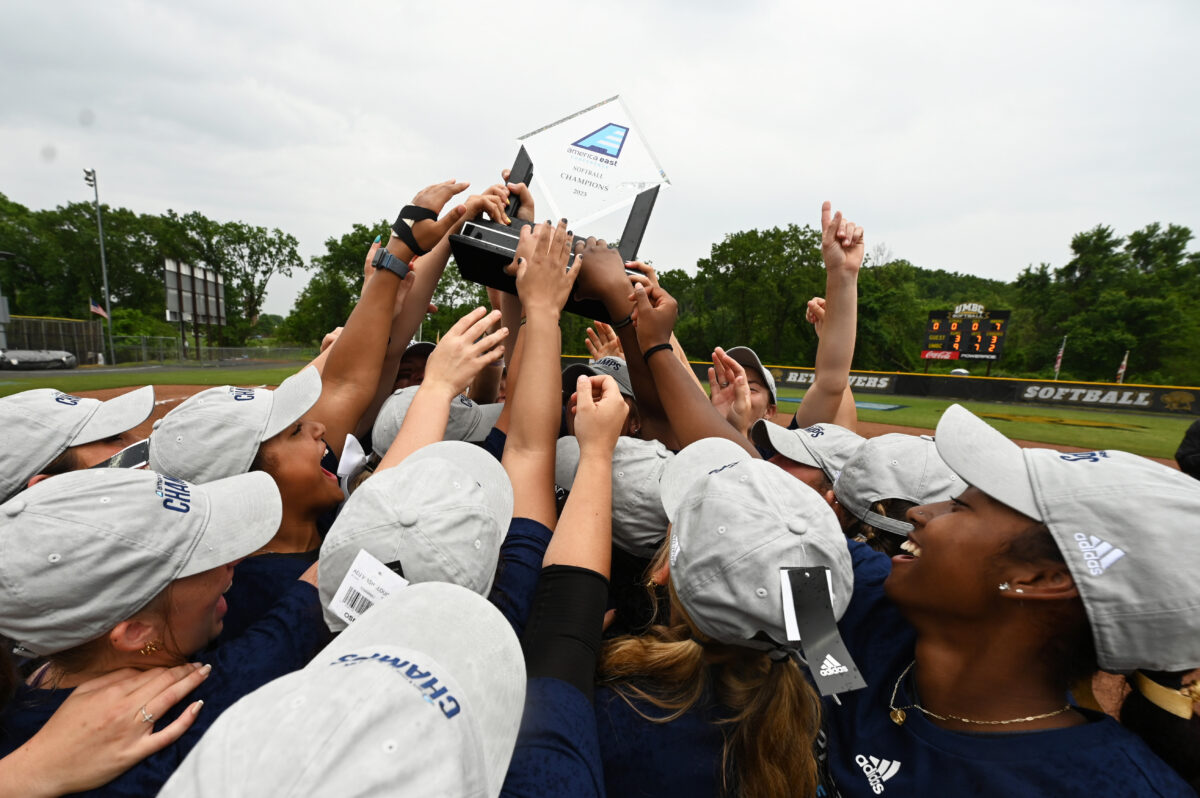 Several hands are raised in the air holding a glass America East trophy on a softball field.