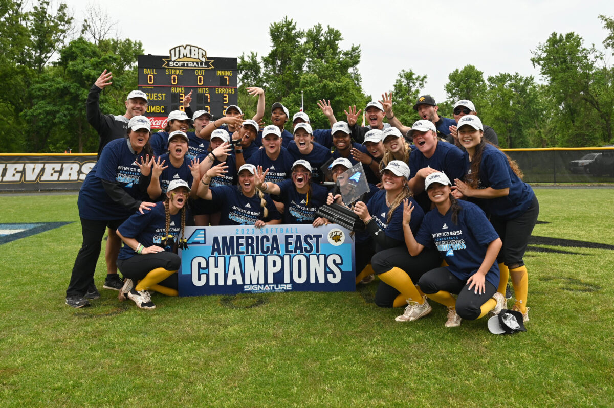 UMBC softball team stands crouched around an America East Champions sign wearing America East hats/