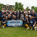 UMBC softball team stands crouched around an America East Champions sign wearing America East hats/