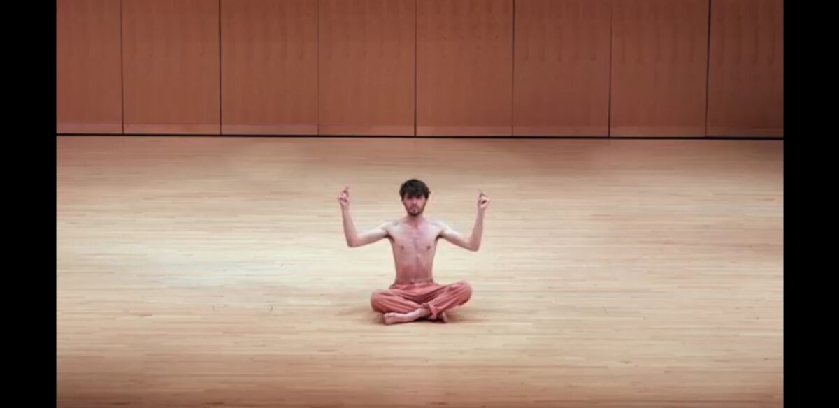 Shirtless man seated on stage performing a contemporary music piece with his body as the instrument. His arms are raised and fingers look to be snapping.