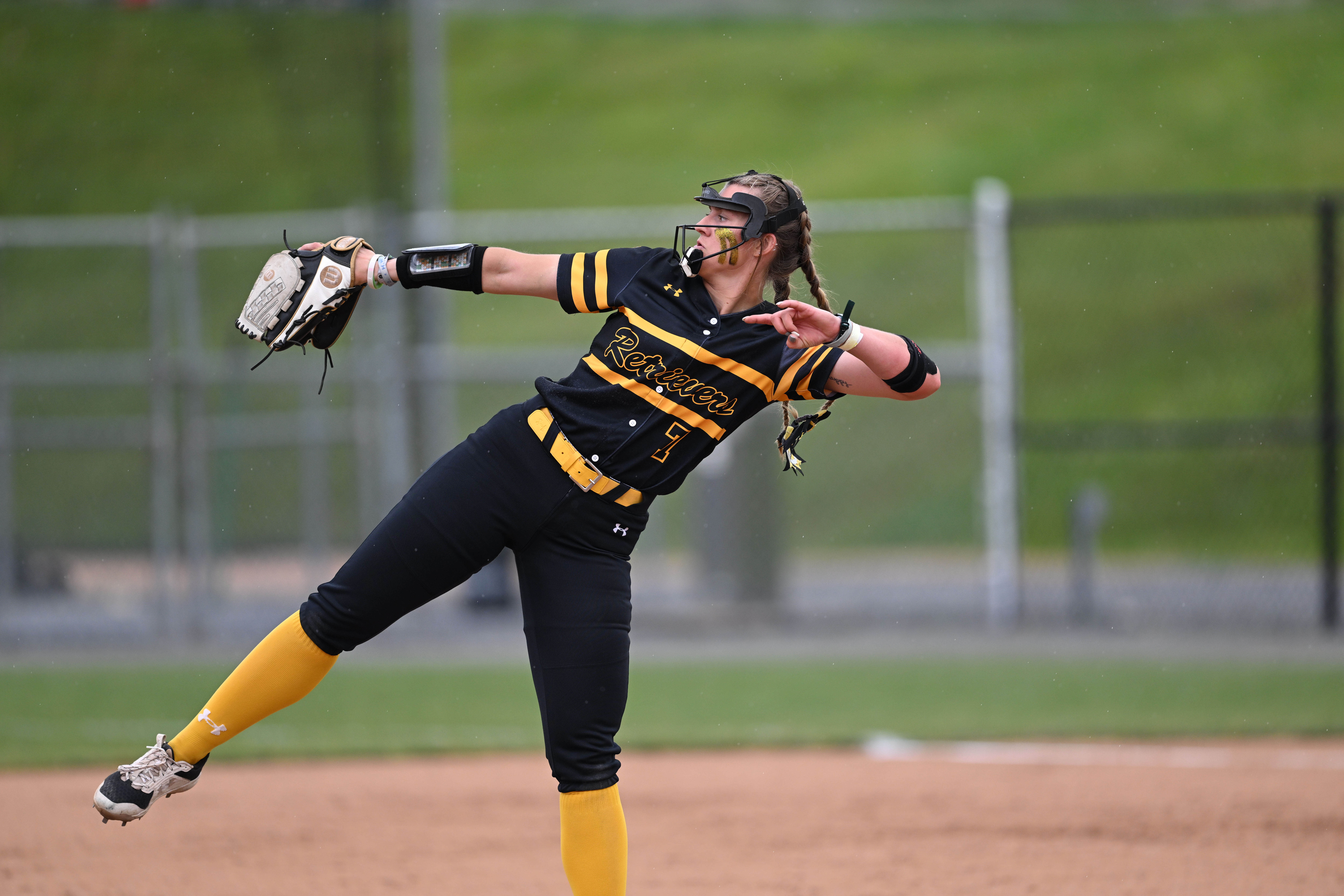 A softball pitcher is reared back on the mound with glove and hand on the same plane wearing a black and gold UMBC uniform.