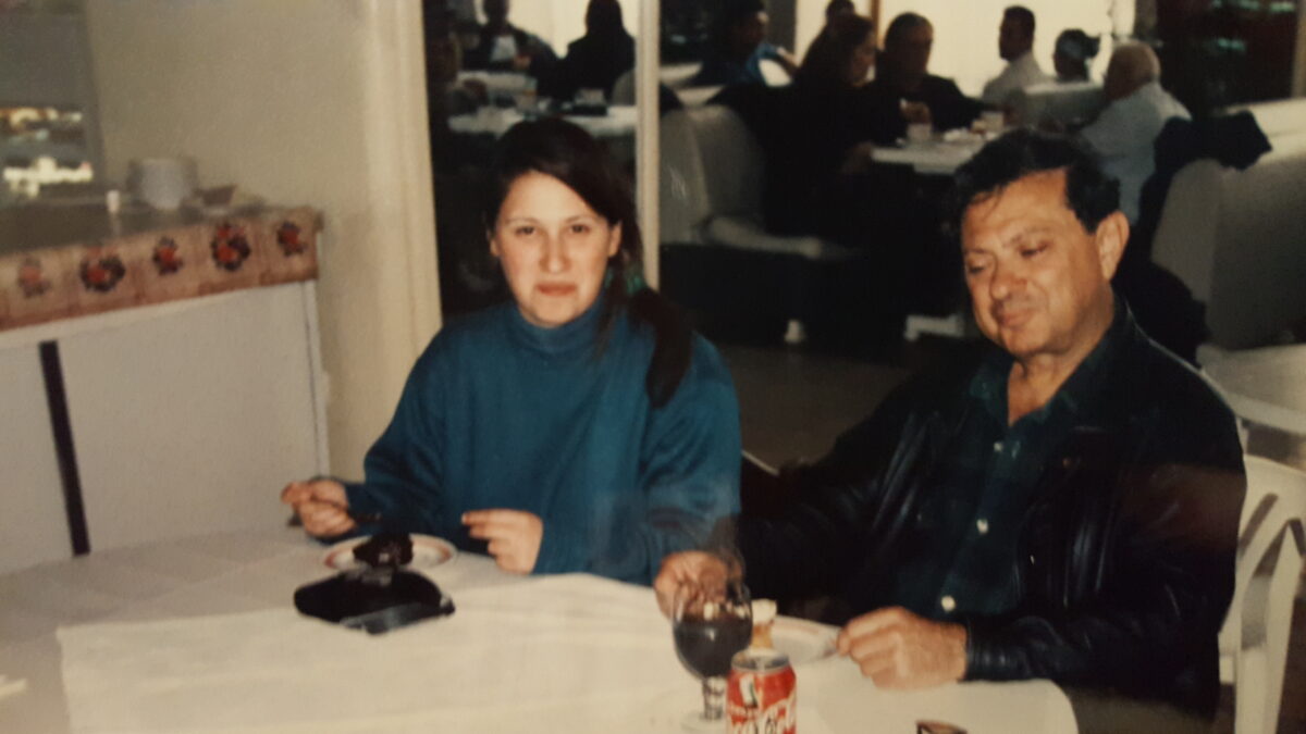 An older photos shows an adult daughter with her father at a restaurant table.