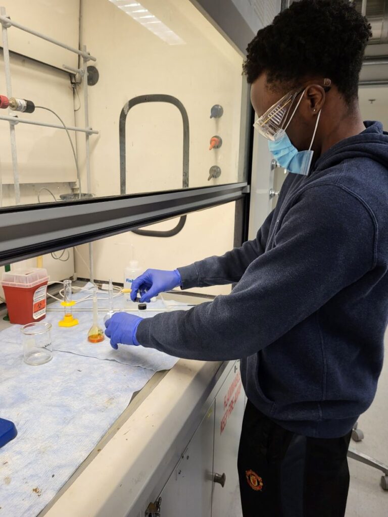 Student wearing protective glasses and gloves works with chemicals under a fume hood.