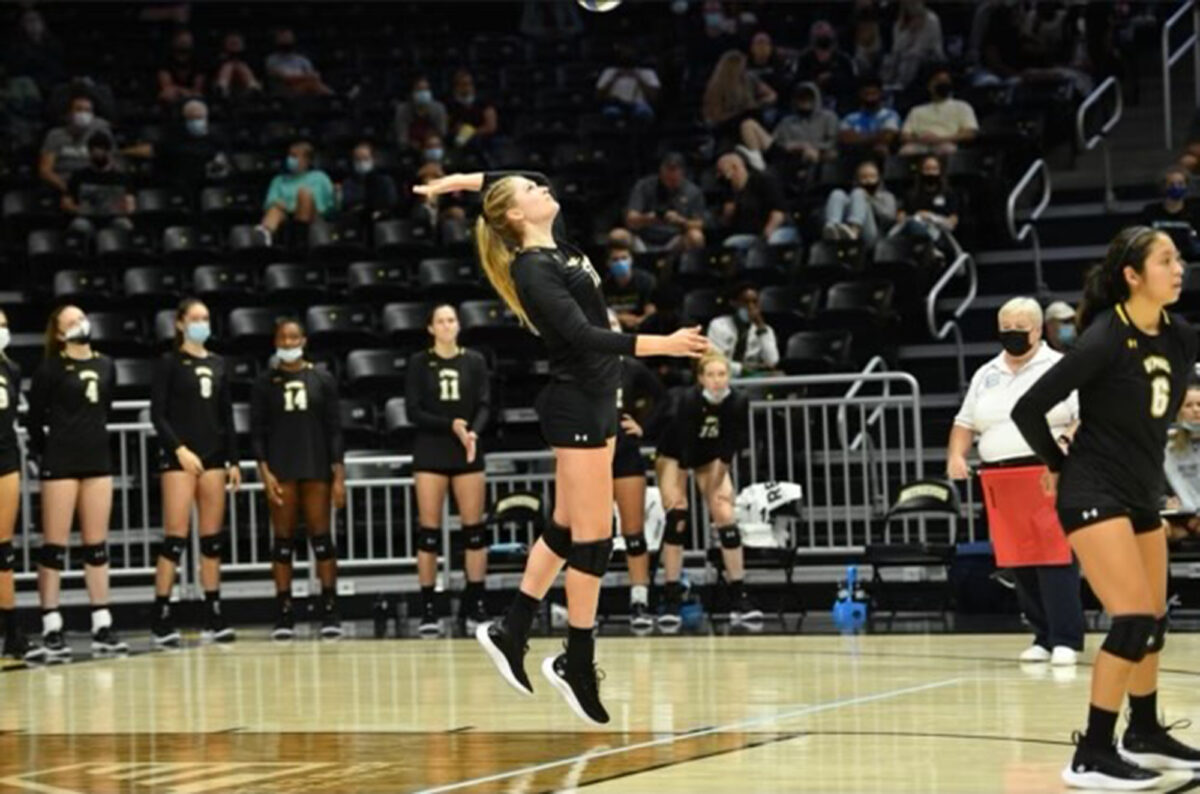 A woman prepares to hit a volleyball on the court. She wears a black uniform with white and gold writing.