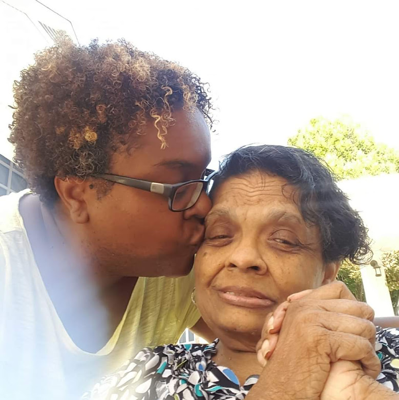 an adult daughter caregiver bends down to kiss her aging mother who has dementia
