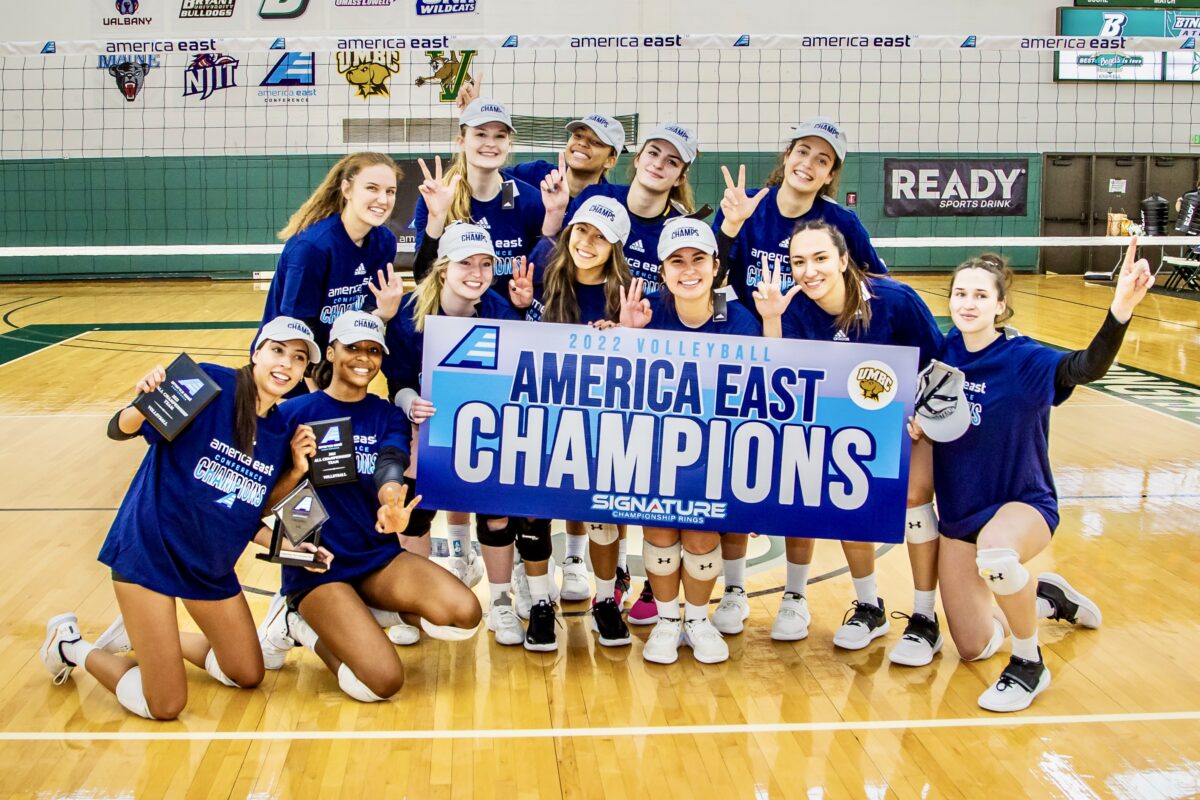 A group of young women pose on a volleyball court with a sign saying America East Champions