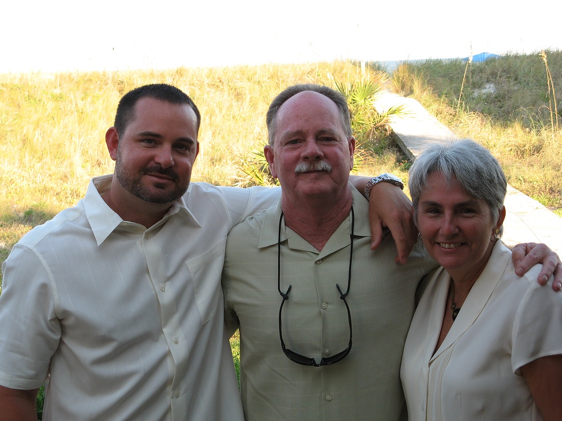 A father and mother pose with their adult son in front of a sandy boardwalk.