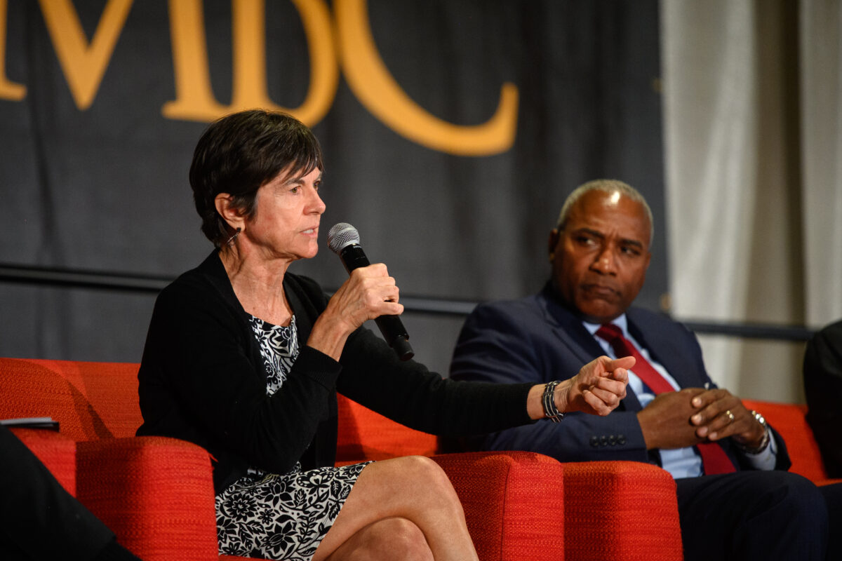 Woman in black and white dress holds a microphone, addressing an audience and sitting on stage next to man in suit and red tie at UMBC-Maryland Matters climate forum.