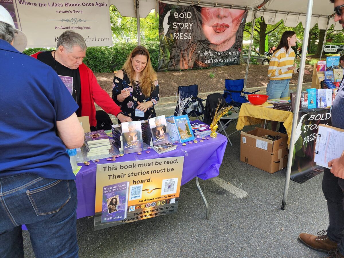 An author stands behind her book table at a book festival. The sign says This book must be read, their stories must be heard.