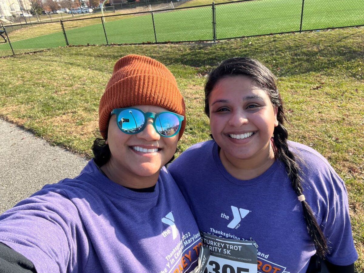 Two women stand in front of grass field and smile at the camera. They wear shirts that read, "The Y" and "Turkey Trop Charity 5K."