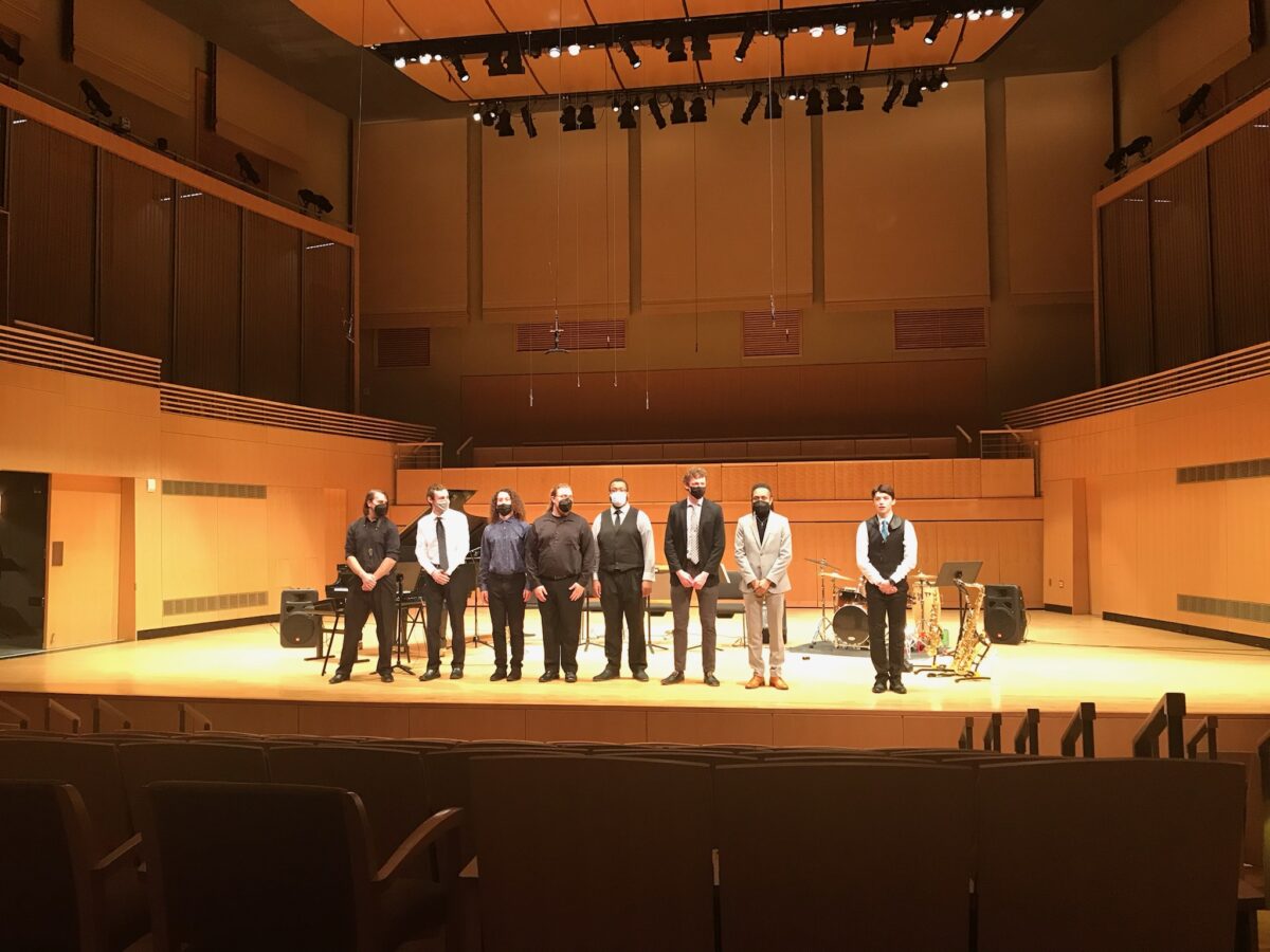 Students standing in a line on stage, in professional attire, with instruments behind them.