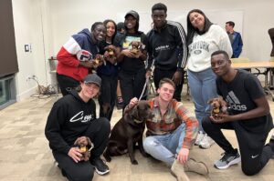 UMBC students (community builders), some standing and some kneeling and sitting, holding a stuffed dog toy. In the center of the photo is a brown dog, Chip, UMBC's campus comfort dog.