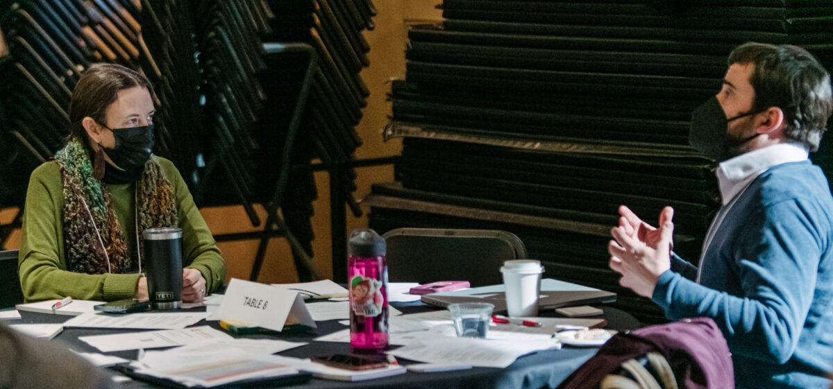 Two colleagues sit at a table filled with papers and cups talking.