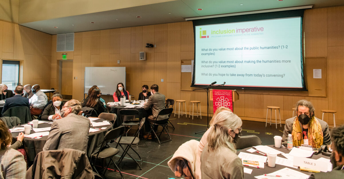 A large room with colleagues seated at round tables with a projection screen of  Inclusion Imperative Convening questions.