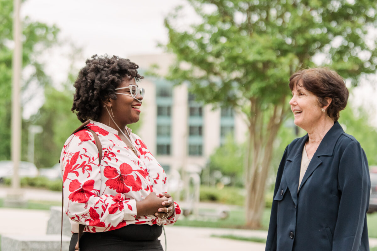 A mentor stands outside talking with a student about asking for help and community support with a building and trees in the background