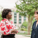 A mentor stands outside talking with a student about asking for help and community support with a building and trees in the background