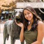 A young woman with dark hair smiles at the camera, posing with a statue of a dog