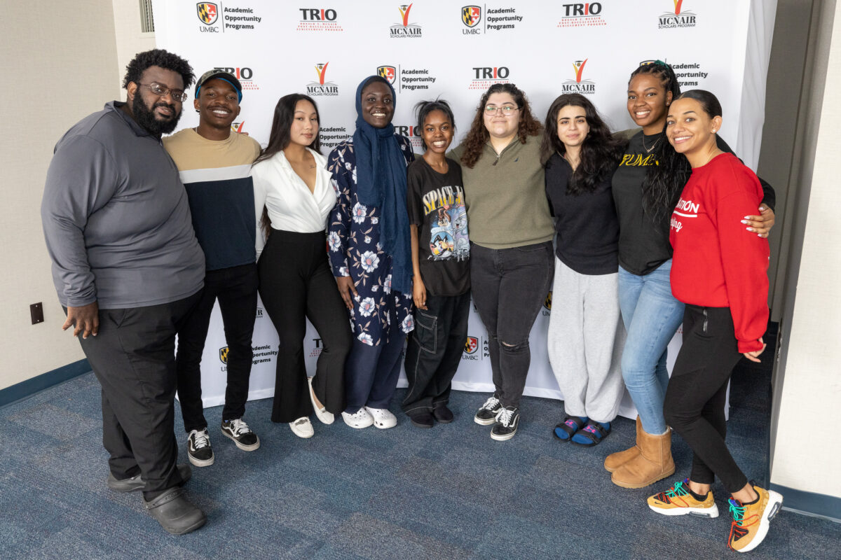 Group of students smiling and posing in front of a backdrop with UMBC Academic Opportunity Programs, TRIO, and McNair Scholar logos.