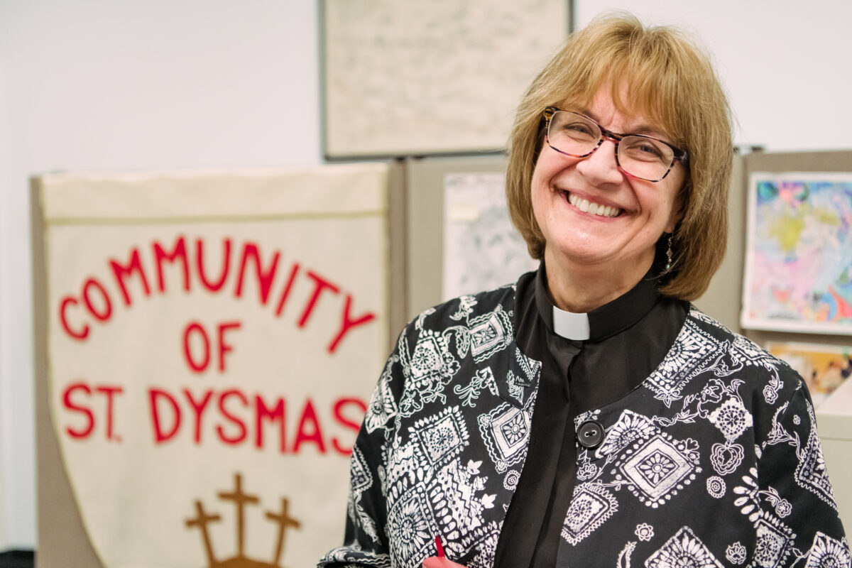 Beck smiles in front of a sign reading, "Community of St. Dysmas."