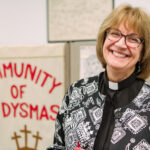 Beck smiles in front of a sign reading, "Community of St. Dysmas."