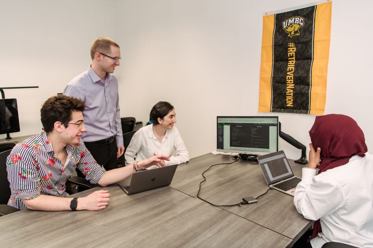 One standing person and three seated people (all AI researchers) look at computer monitor placed on a table. A banner on the wall reads "UMBC" and "#RetrieverNation"