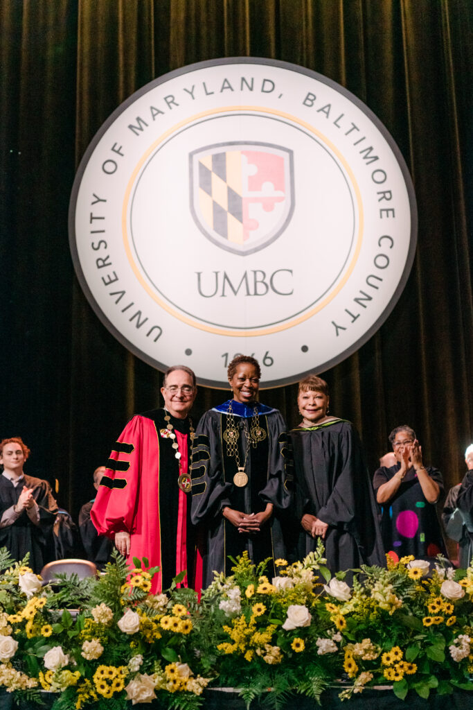 Three individuals in regalia stand behind of a display of flowers and in front of the UMBC seal