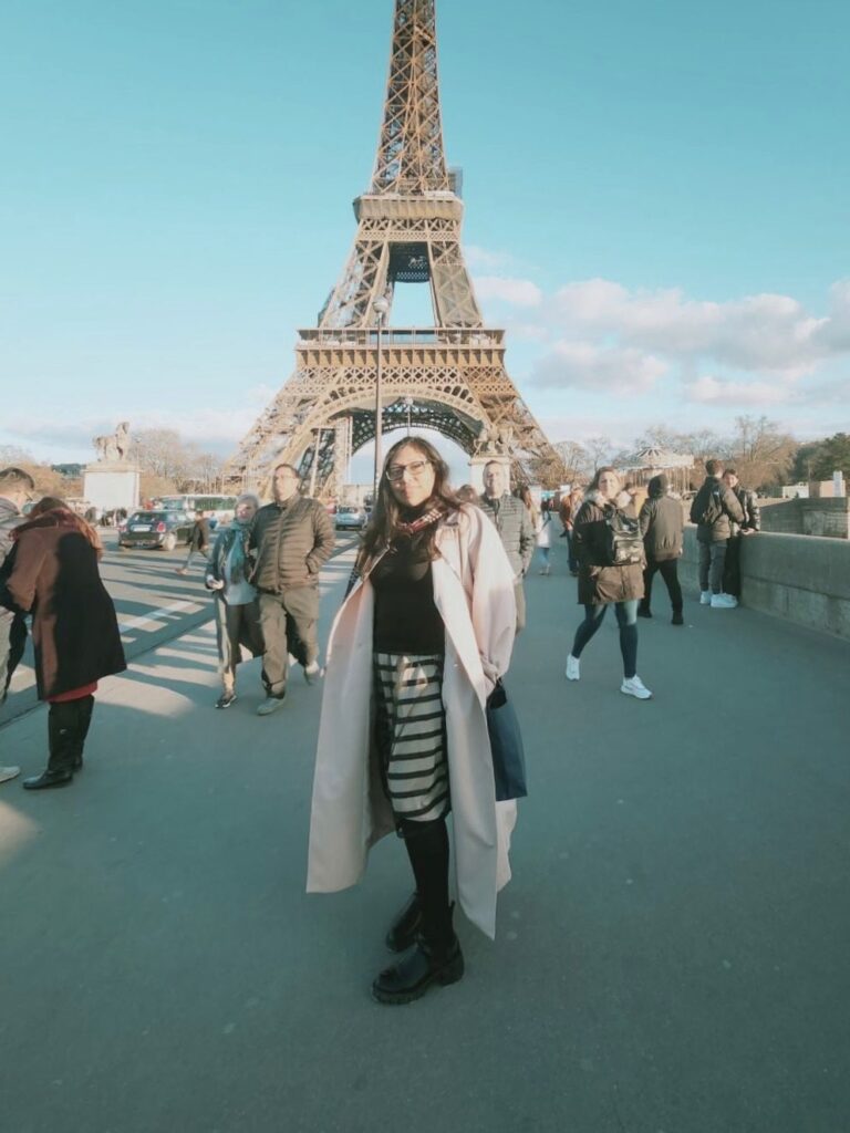 A person with a long trench coat stands in front of the Eiffel Tower