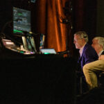 A man sits in a production booth with computer monitors showing him information