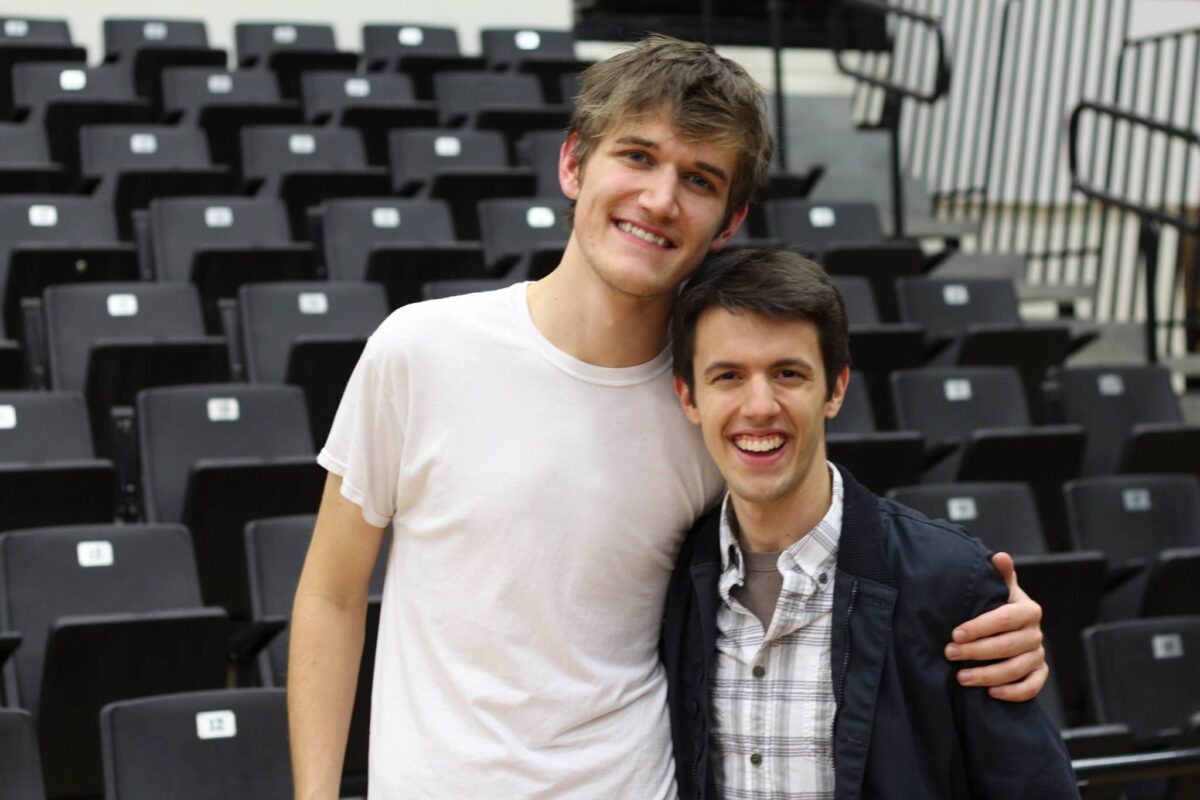 two guys stand side hugging in front of stadium bleachers