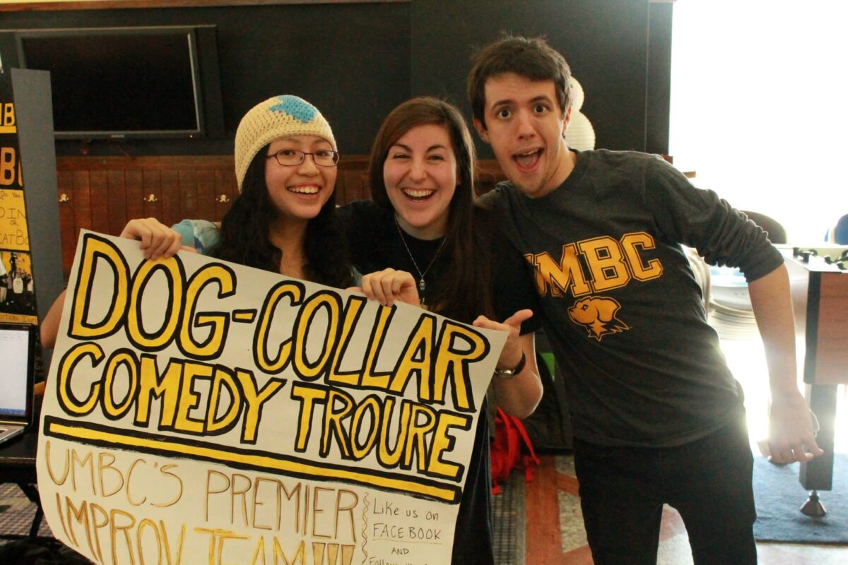 three people stand smiling together with a sign that says Dog Collar Comedy Troupe