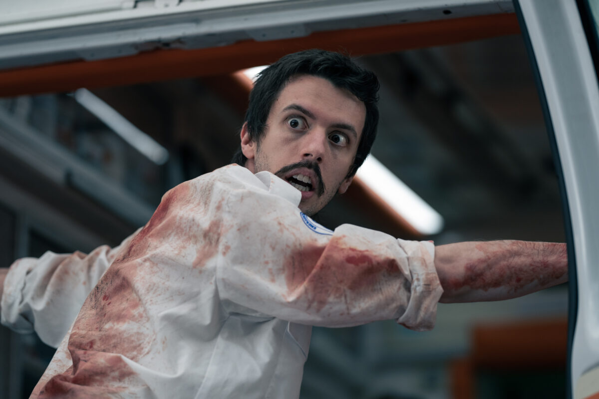 in a still from the Cocaine Bear movie, a paramedic turns around from an ambulance, blood is on his white shirt