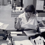in a black and white photo, a woman sits at a desk, pouring over materials