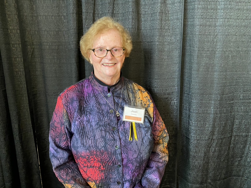 a headshot of Joan Costello--a Retriever for life--a woman in a bright top against a black backdrop