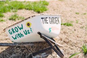 an unused, decorative wheel barrow lays on its side. Painted on the side says The Garden, grow with us!