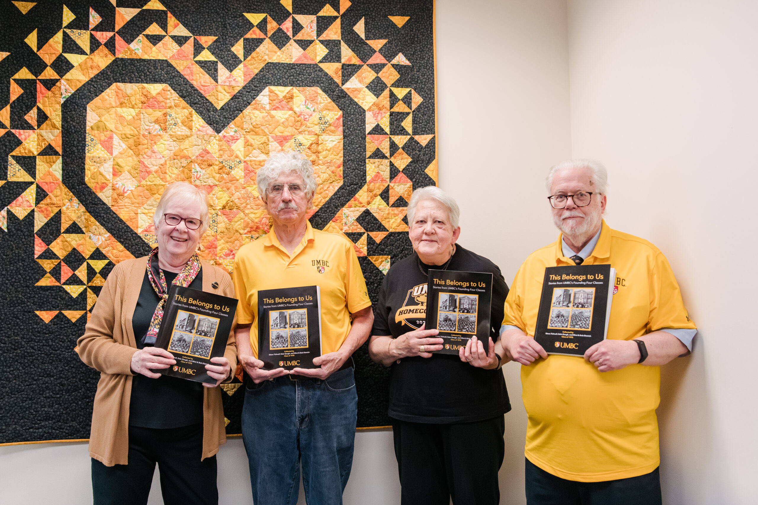 Four people stand in front of a quilt with a gold heart on it. The people are holding copies of a book titled This Belongs to Us.