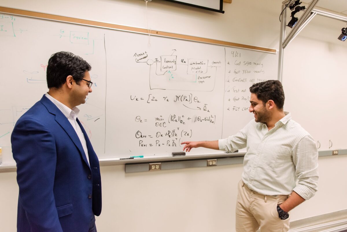 One man points to equations on a white board and discusses with another man.
