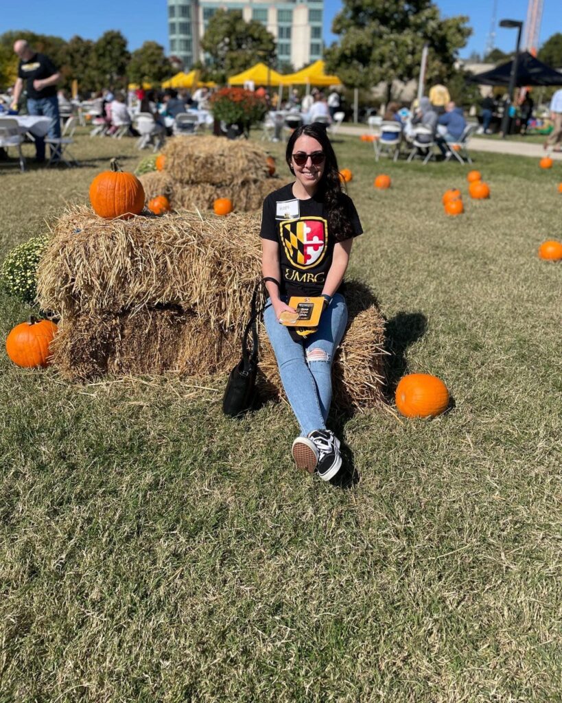 A woman in sunglasses sits on a haybale surrounded by pumpkins.