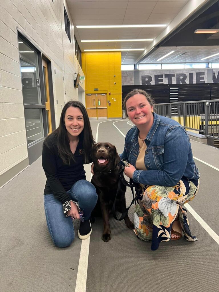 Two women kneel next to a brown dog. Both help transfer students succeed at UMBC.