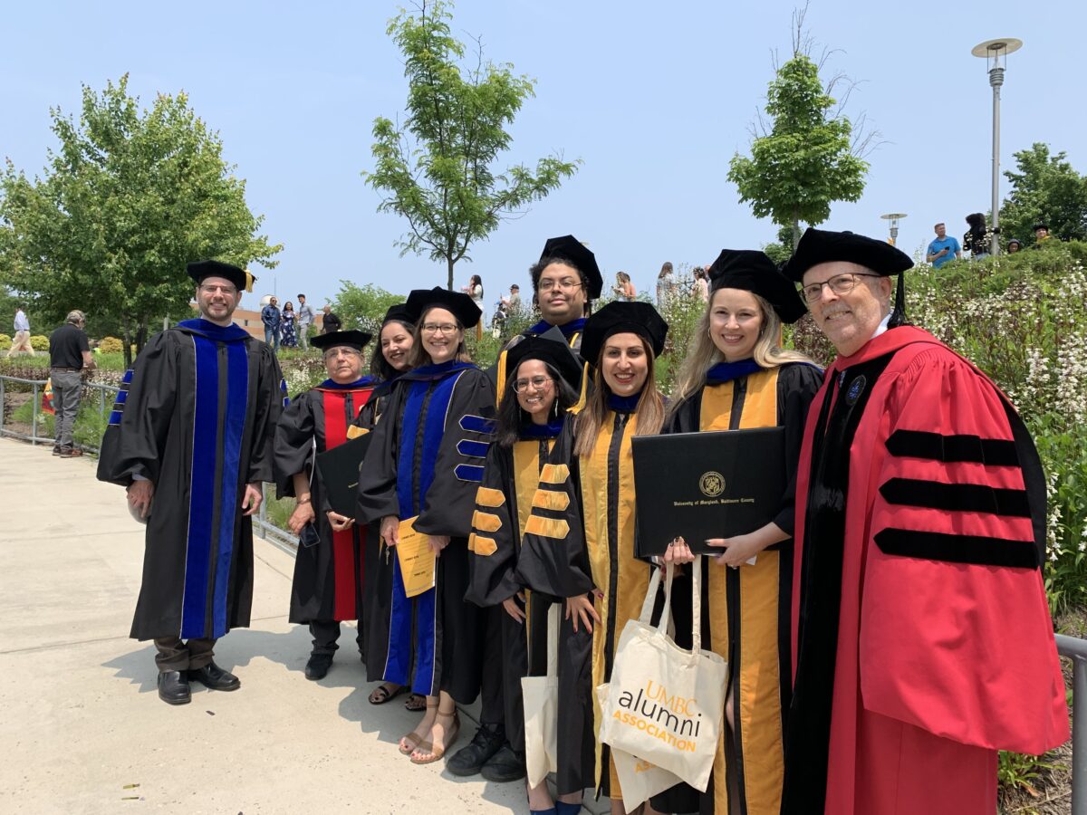 group photo of nine people in graduation regalia outdoors on a sunny day