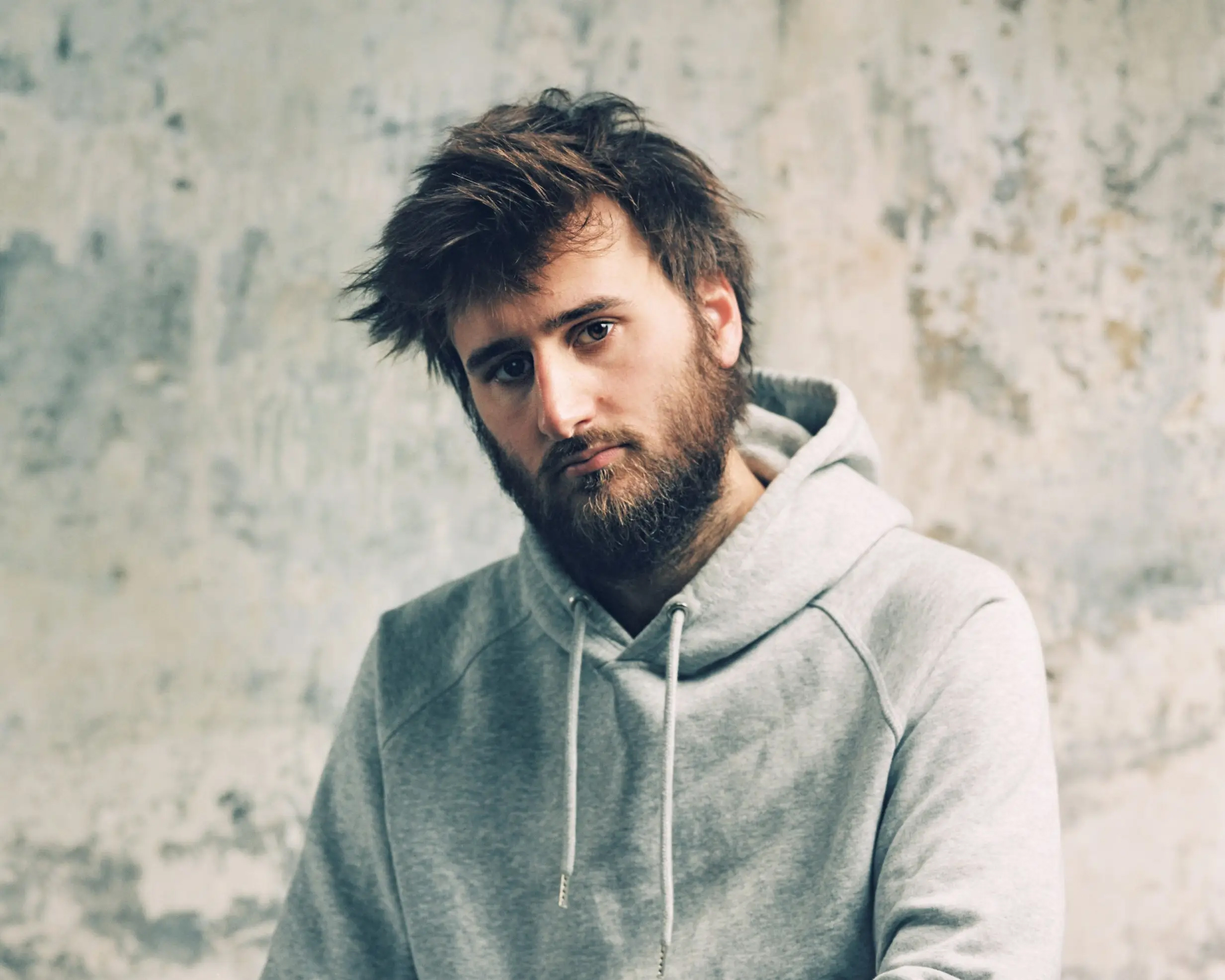 A man with dark hair and a beard, wearing a sweatshirt, looks at the camera