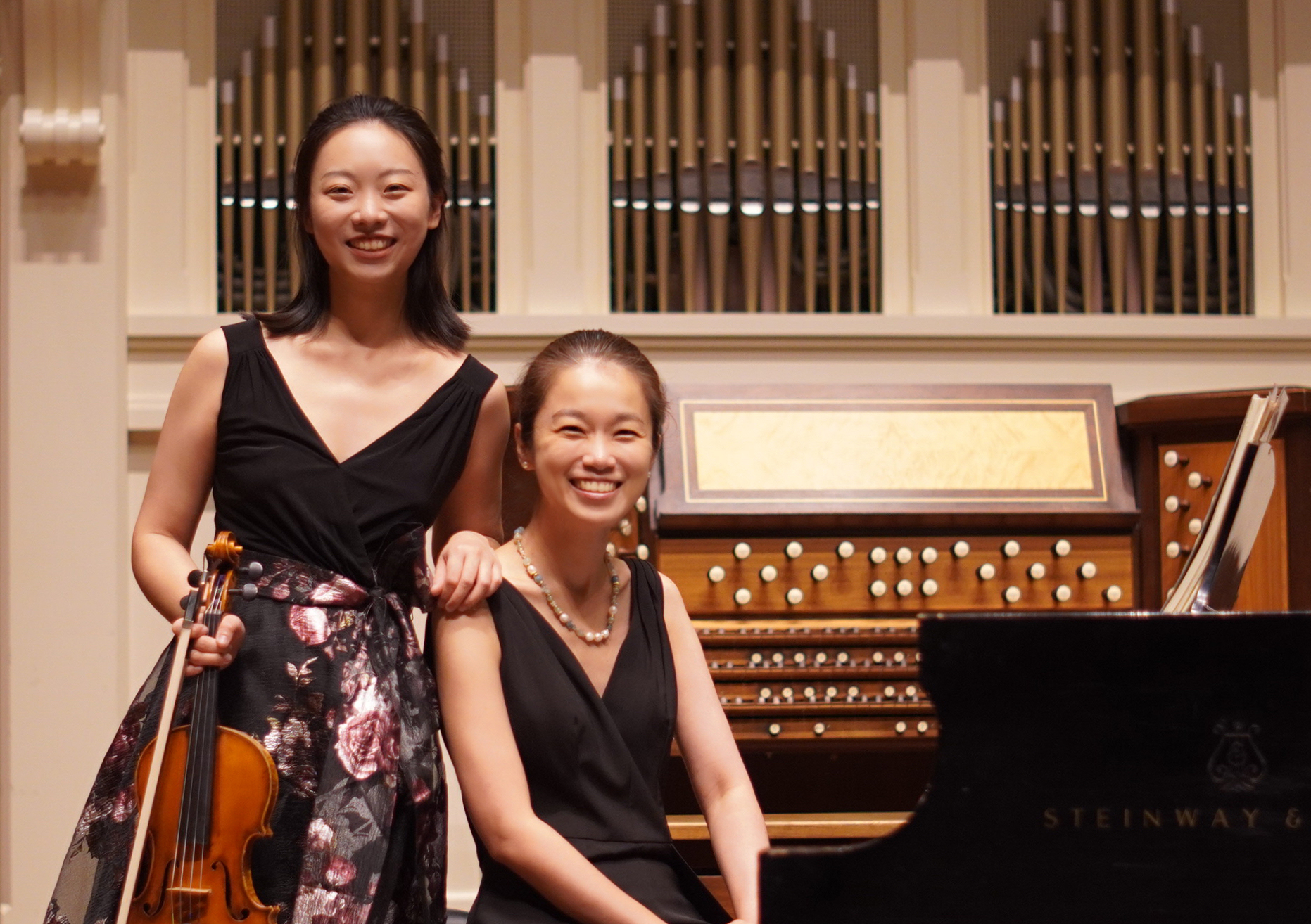 Two musicians, one with a violin and one at the piano, smile at the camera