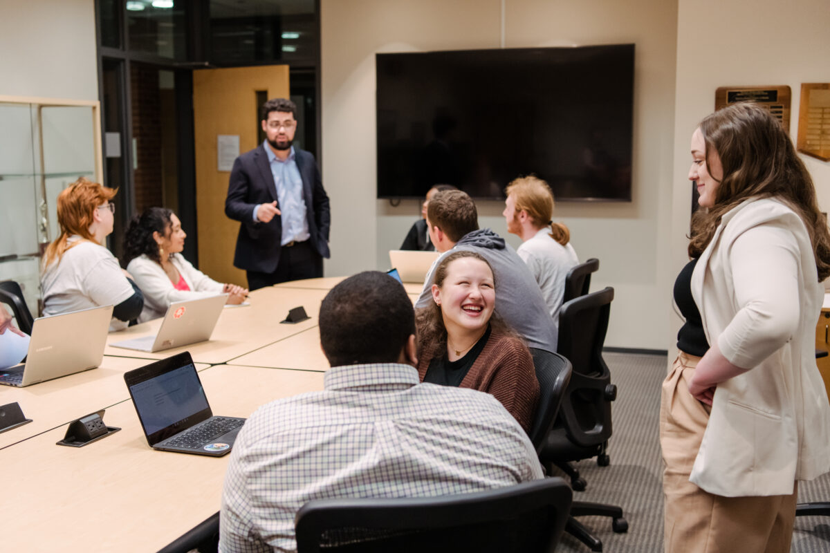 a group of students sit around a conference table, looking up as alumni speak to them