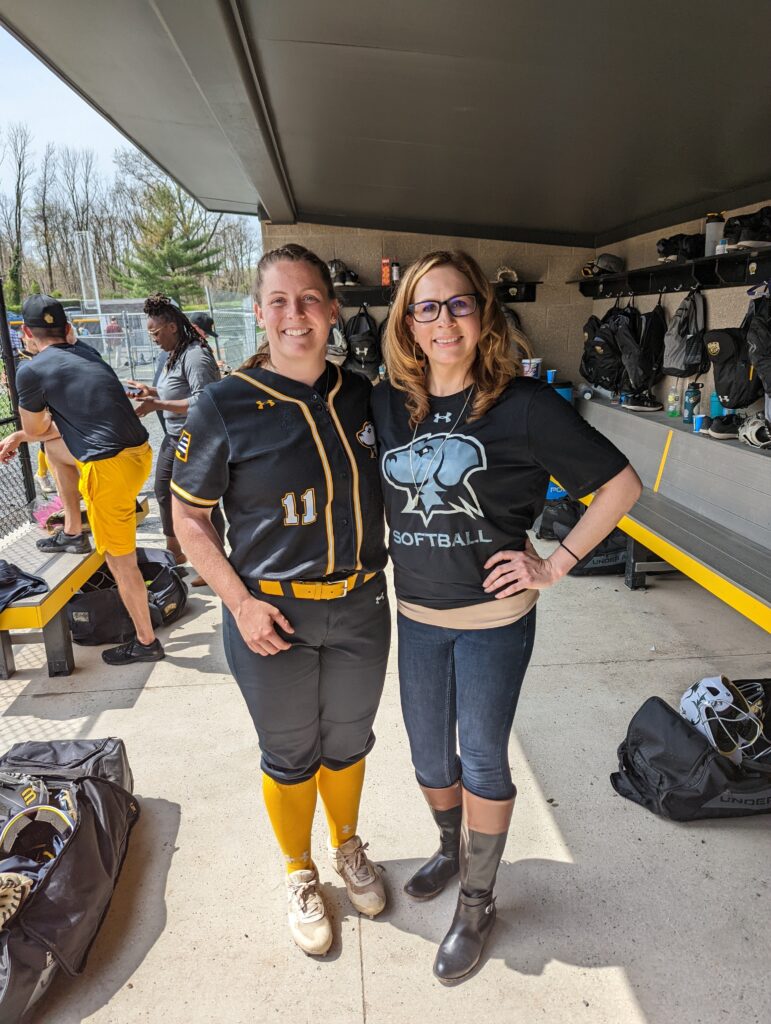 A softball player stands in a dugout with an honorary faculty coach