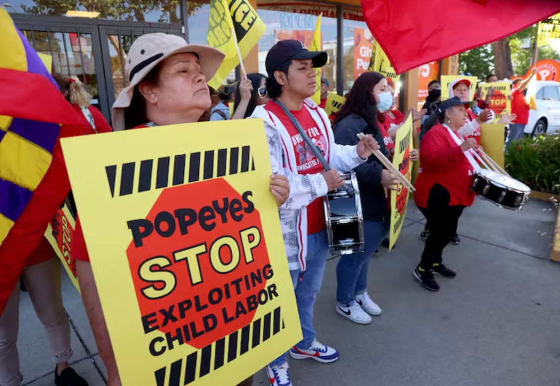 Workers protest outside a Popeye’s restaurant in Oakland, Calif., on May 18, 2023. Workers are holding a "Popeyes stop exploiting child labor" sign" along with participants holding instruments like drums. 