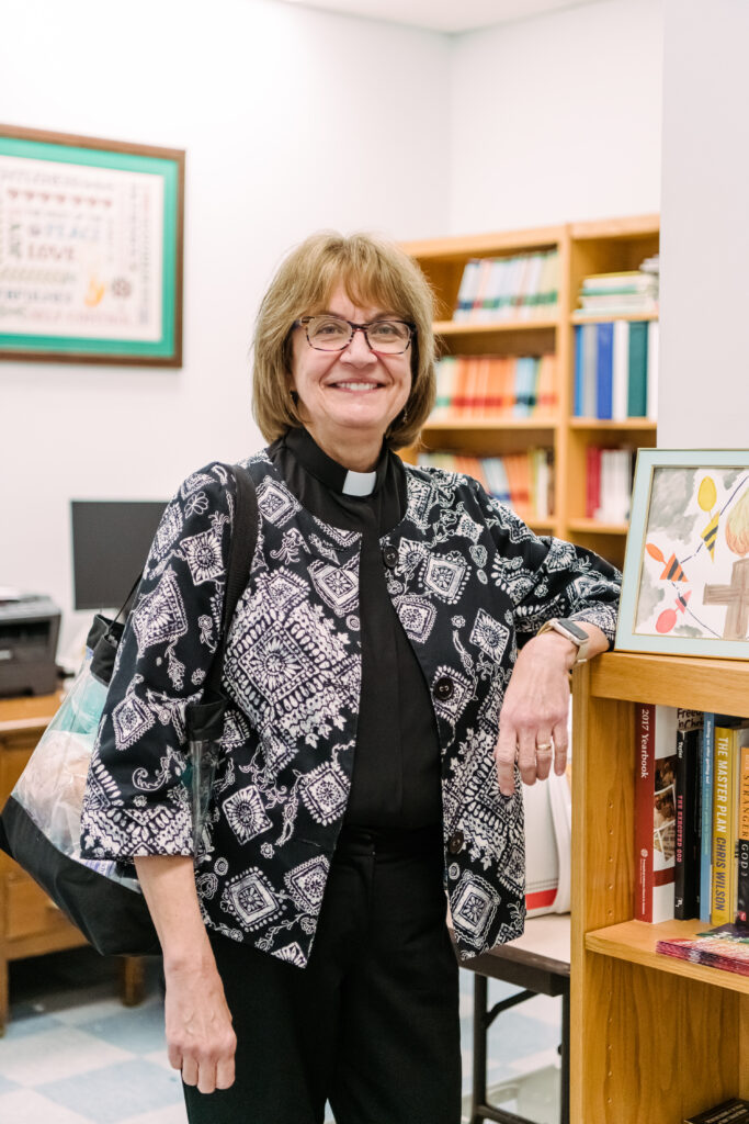 Reverand Susan Beck in her donated office space where she prepares materials for ministering to her prison congregation in Maryland. She stands, smiling, with her bag, ready to go.