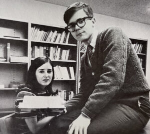 black and white photo of a young man and woman sitting at a desk with a bookcase behind them