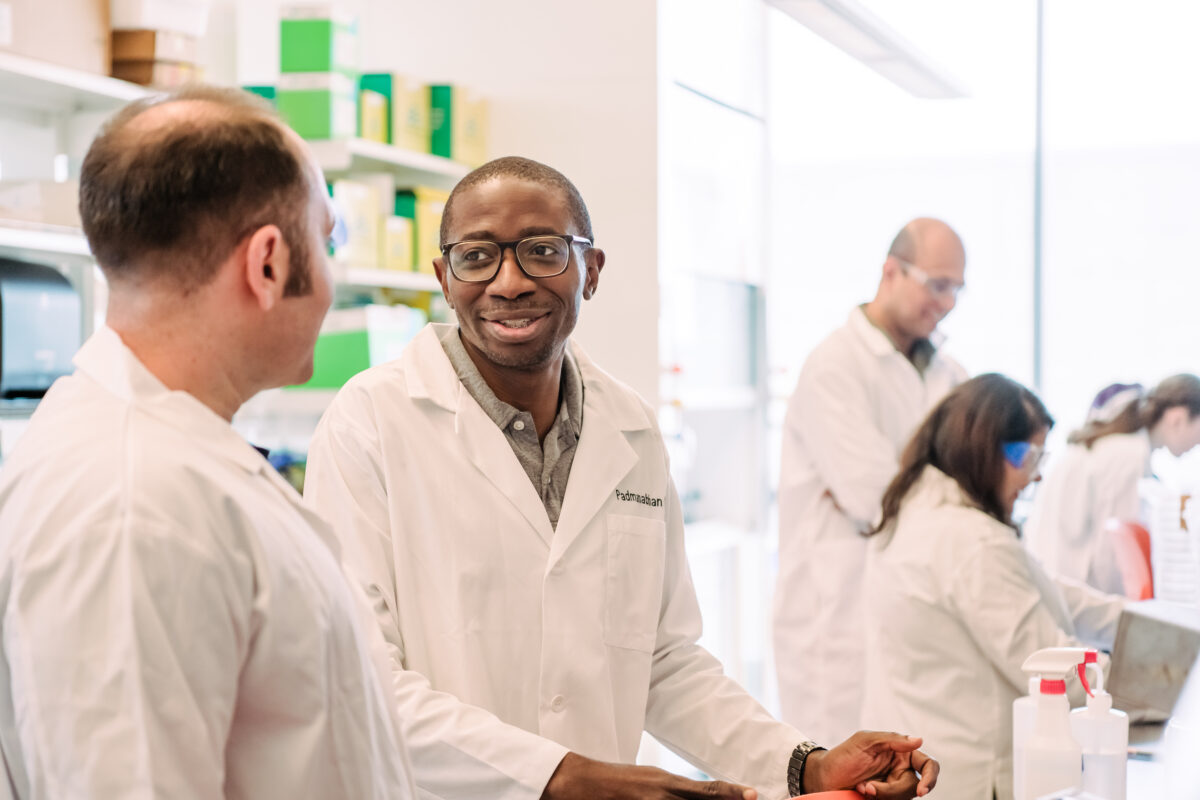 two people wearing lab coats converse in a brightly lit laboratory; others are at work at a lab bench in the background